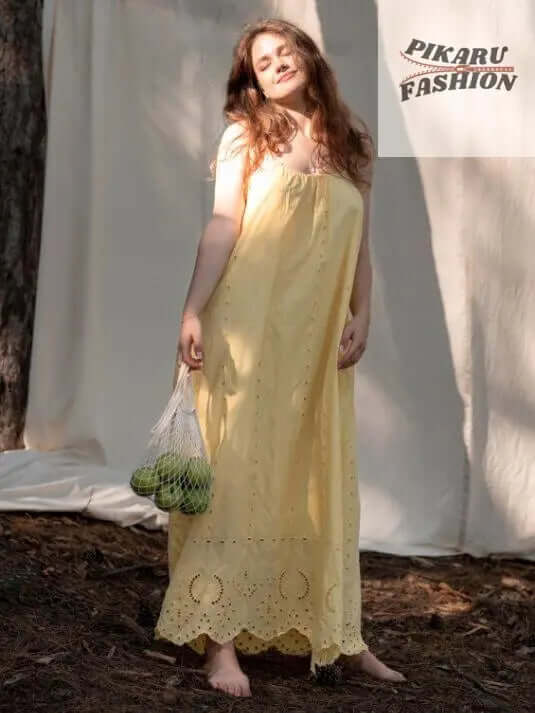 Woman in yellow boho dress holding apples in mesh bag, standing outdoors by tree.